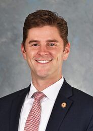 Smiling man in a dark suit and pink tie against a gray background.