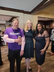 Three women pose indoors, wearing dark dresses and a purple shirt, smiling at an event.