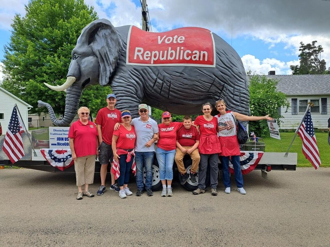 Group in red shirts posing before a giant elephant sign reading “Vote Republican” at an outdoor campaign event