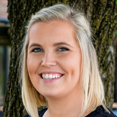 A woman with blonde hair is smiling in front of a tree.