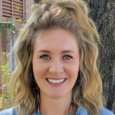 A woman with blonde hair is smiling for the camera in front of a tree.