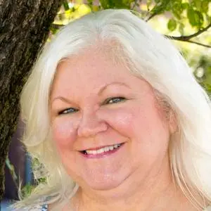 A woman with white hair is smiling in front of a tree.