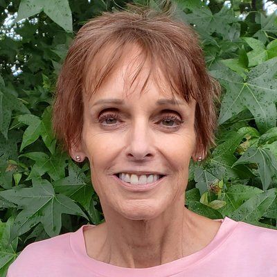 A woman in a pink shirt is smiling in front of a bush.