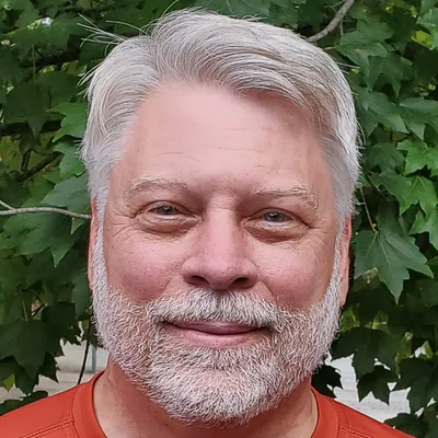 A man with gray hair and a beard is smiling in front of a tree.