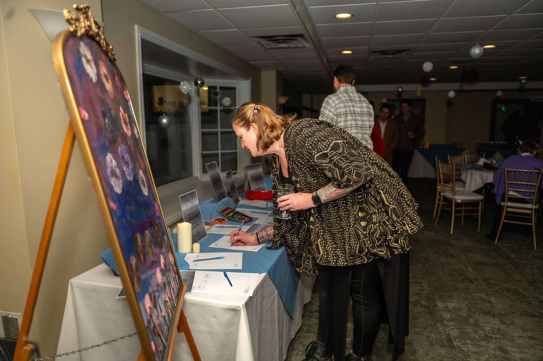 Woman signing a paper at a registration table, framed artwork on display, event in a room.