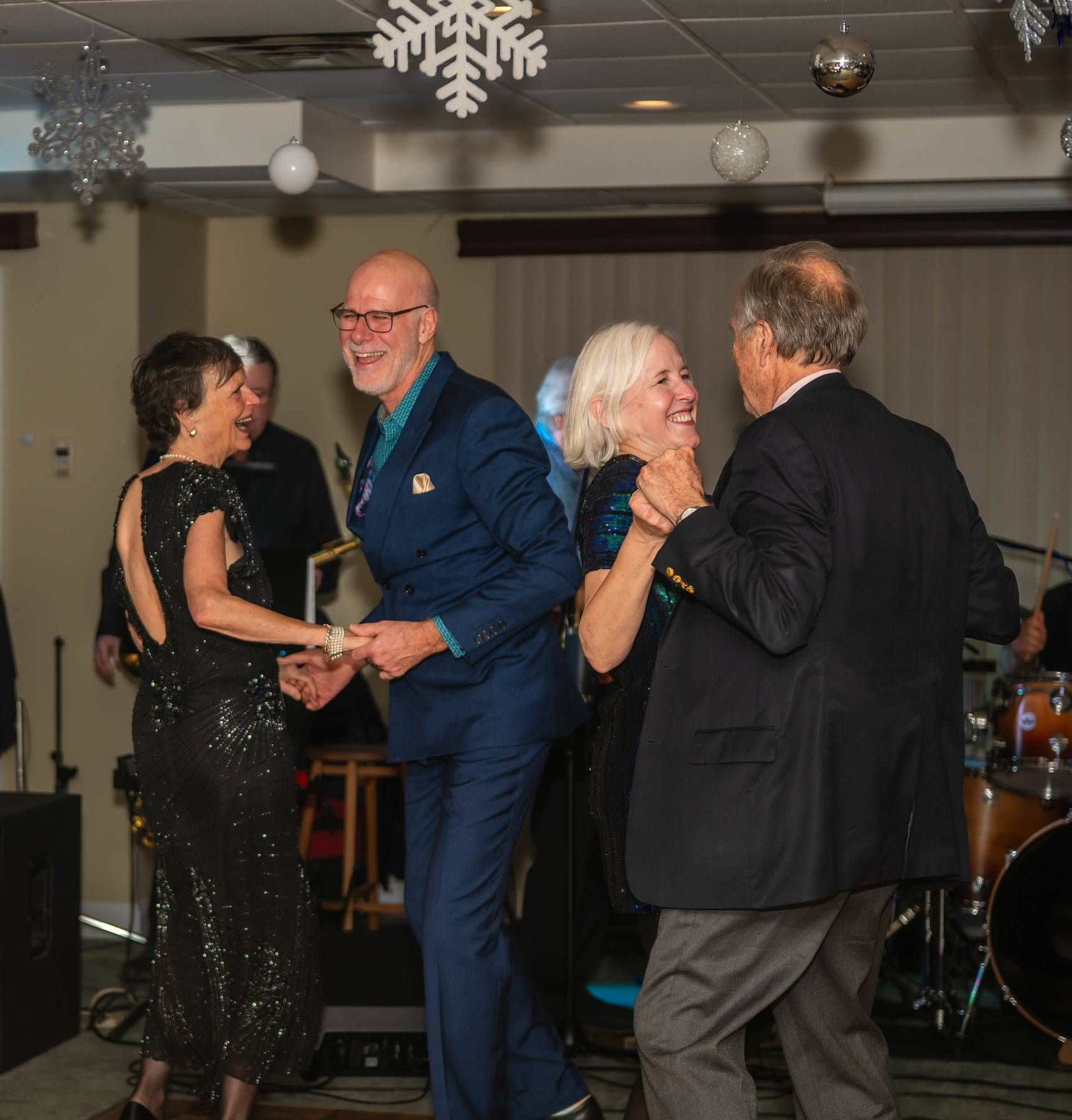 Two couples dancing at a formal event, with a band in the background and festive decorations.
