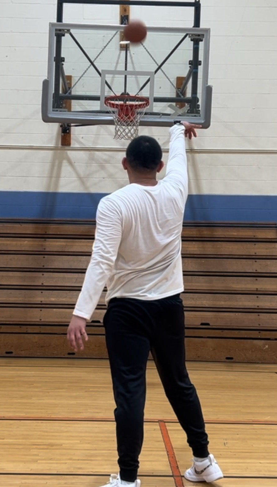 A person in a white long-sleeve shirt and black pants reaches up to touch a basketball backboard in a gym.