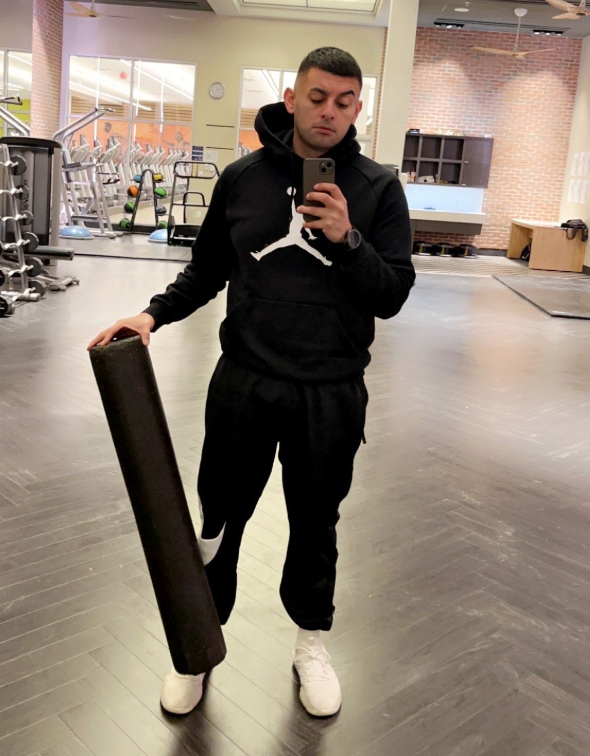 A person stands in a gym, wearing a black Jordan hoodie and sweatpants, holding a foam roller while taking a mirror selfie.