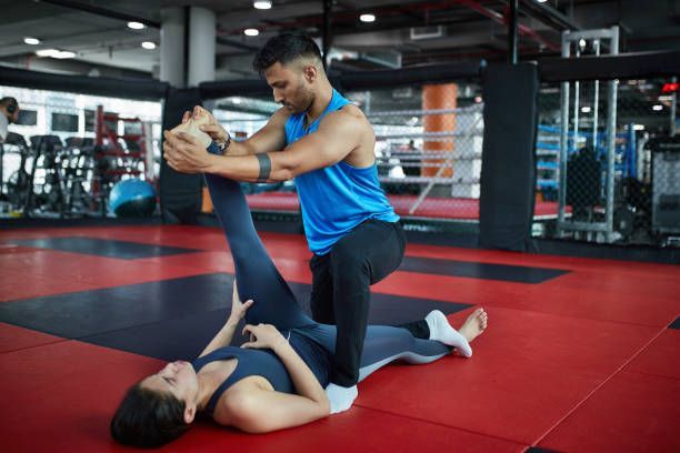 A trainer helps a person with a leg stretch while lying on a gym mat in a fitness studio.
