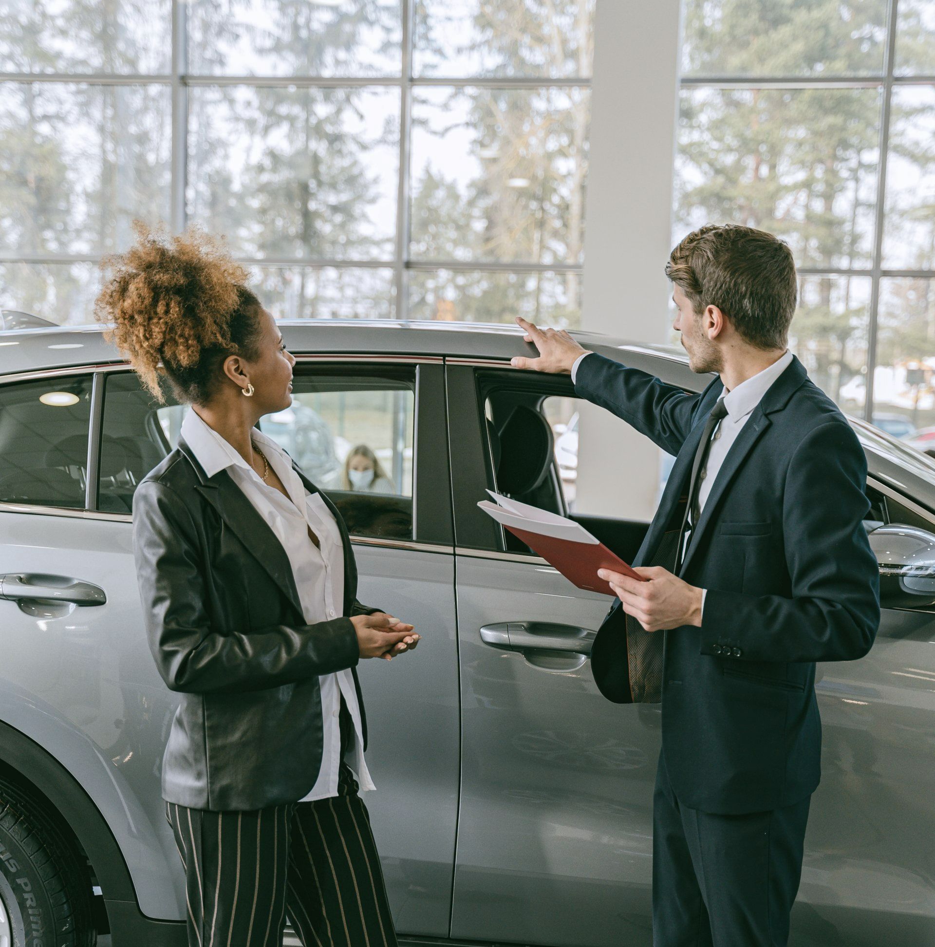 A car salesperson in a suit points to the roof of a gray car while talking to a customer in a dealership showroom.