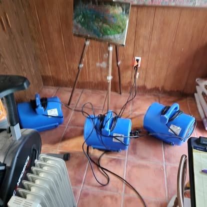 Three blue electric fans on a tiled floor facing a landscape painting on a wooden wall