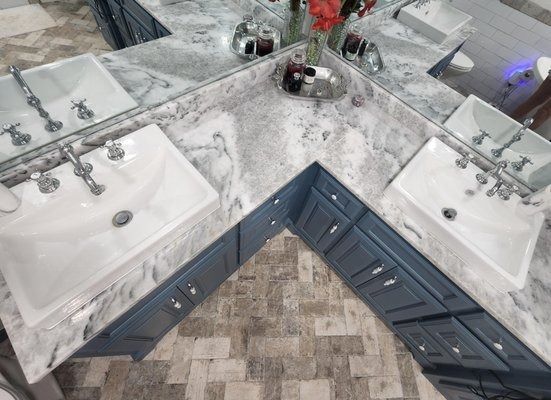 Corner bathroom vanity with two white sinks, marble countertop, and blue cabinets above mosaic tile floor