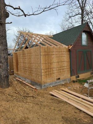 Partially built wooden shed with exposed roof framing beside a red barn in a rural yard