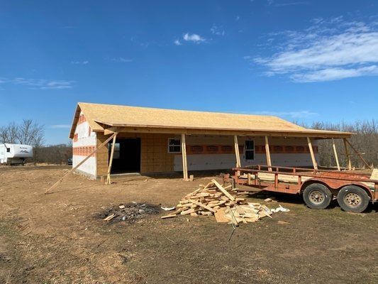 Partially built wooden house with a trailer and construction debris in a rural field under a blue sky