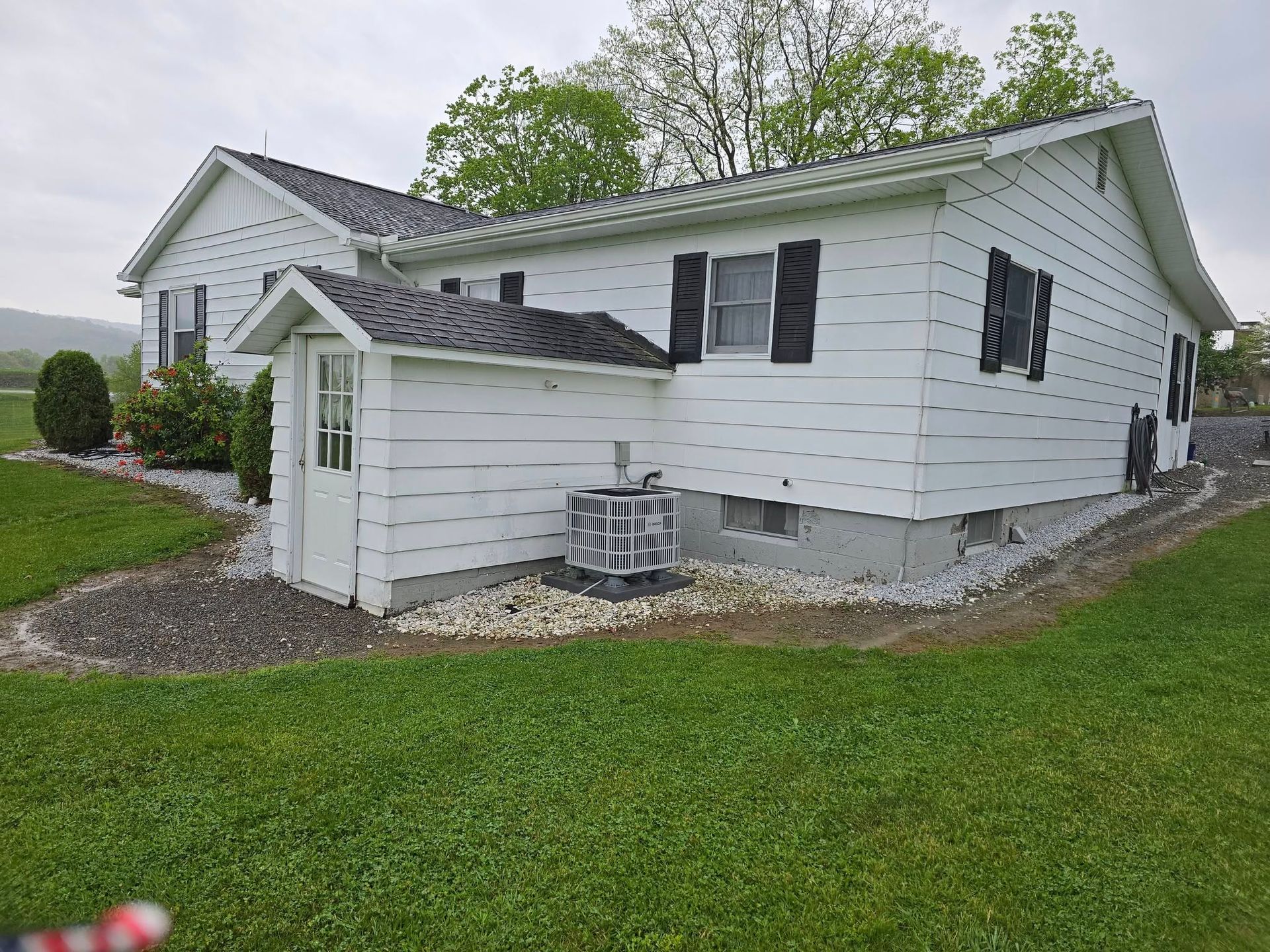 A white house with black shutters, a small shed-like entryway, and an HVAC unit on a gravel perimeter in a grassy yard.
