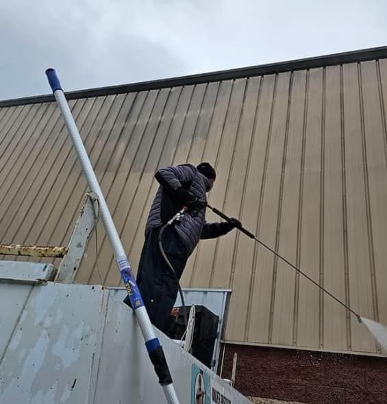 A person in dark clothing uses a long-reach pressure washer to clean a tall, tan-colored metal building wall.
