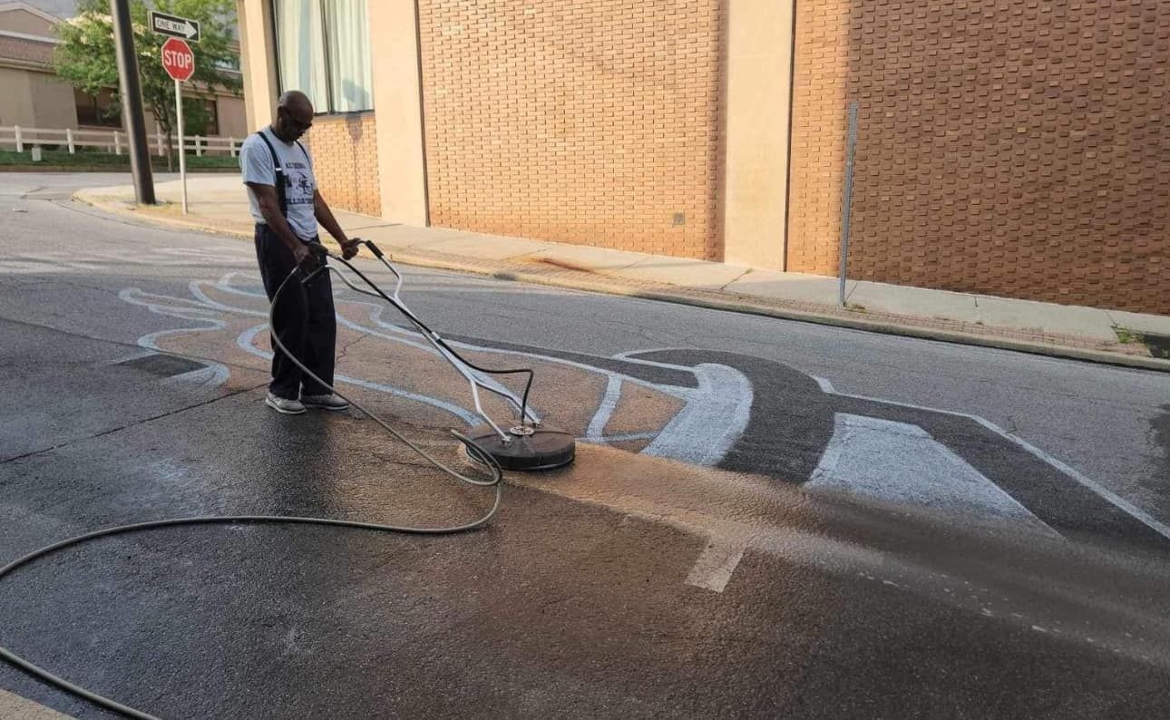 A person cleans a dirty street surface using a circular pressure washer near a brick building.