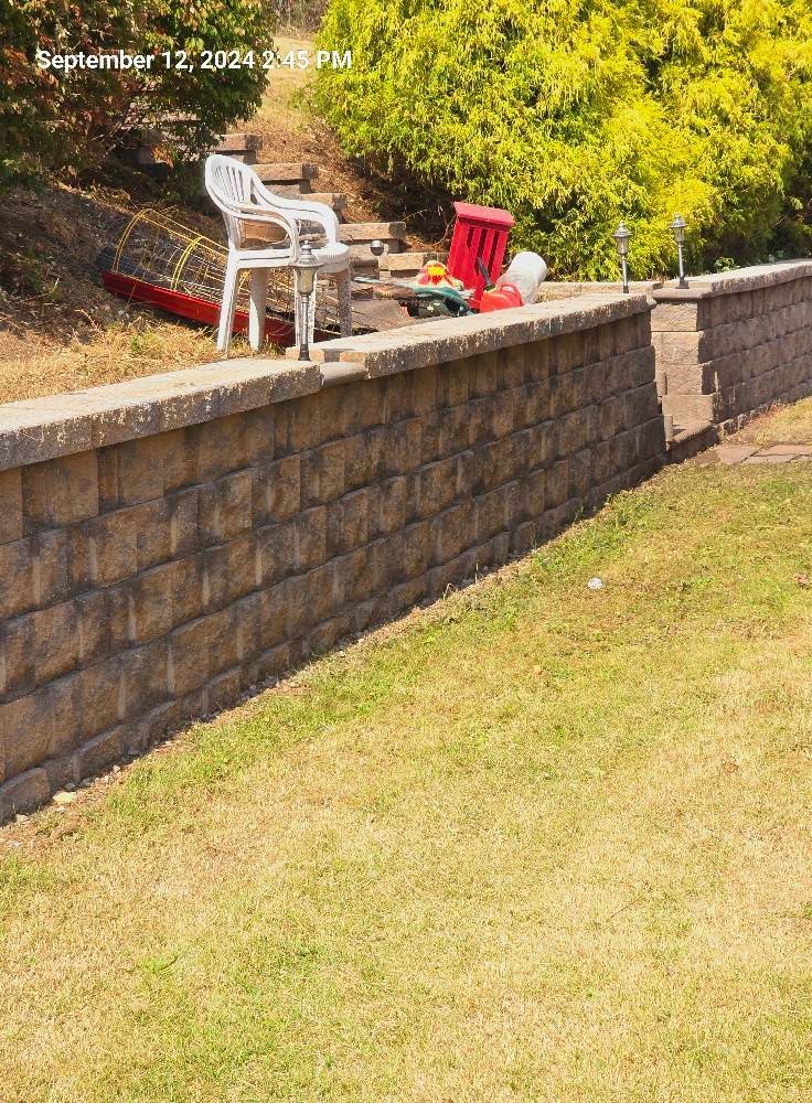 A white patio chair and red equipment sit on a grassy hill behind a stone retaining wall in a sunny outdoor setting.