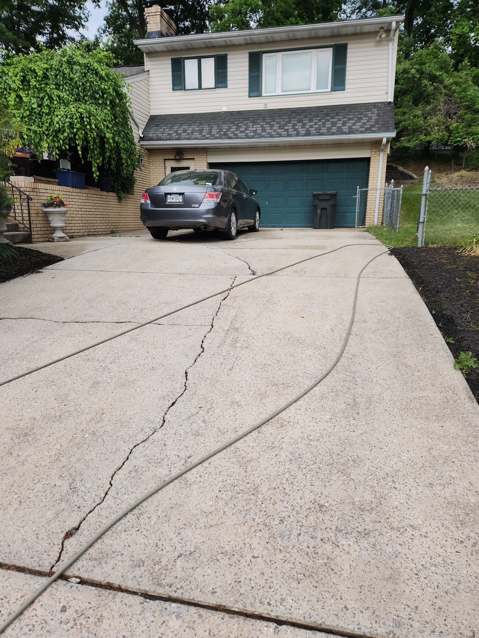 A house with a dark green garage door, a gray sedan parked in the driveway, and a visible crack in the concrete.