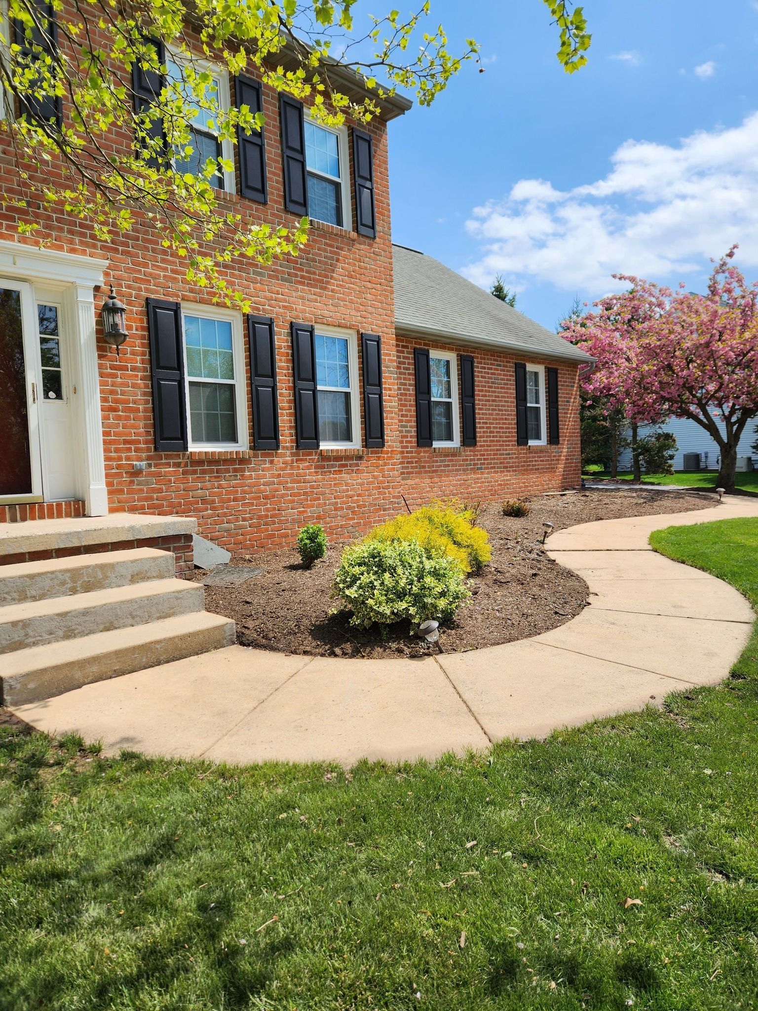 A two-story red brick house with black shutters, concrete steps, and a curved walkway beside a blooming pink cherry tree.