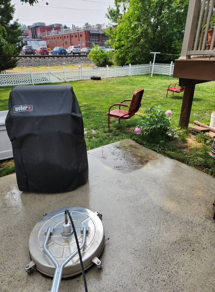 A pressure washer surface cleaner sits on a concrete patio next to a covered grill in a grassy backyard.
