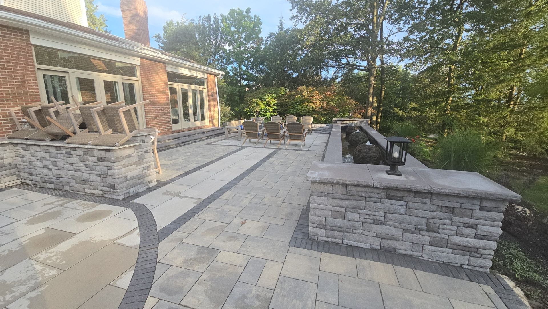 A patio with stone pavers, a brick home, a stone retaining wall, and outdoor furniture under trees.