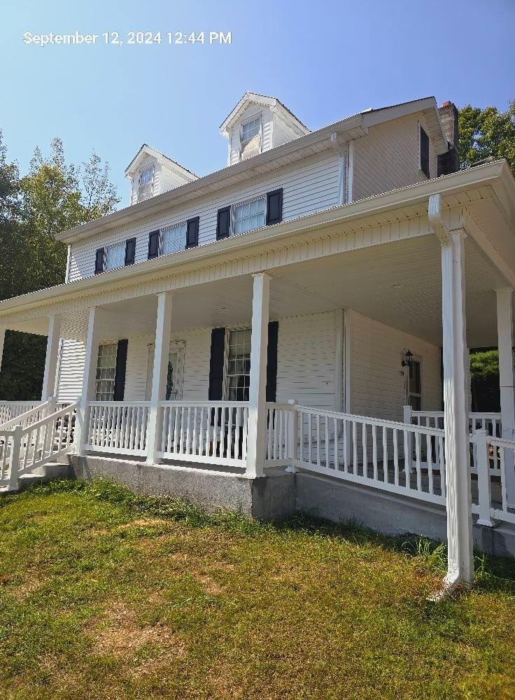 A two-story white house with a large wraparound porch, dark shutters, two roof dormers, and a grassy front yard.