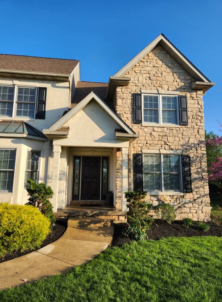 A two-story suburban home with tan stone siding, a covered entryway, black shutters, and a front walkway on a sunny day.