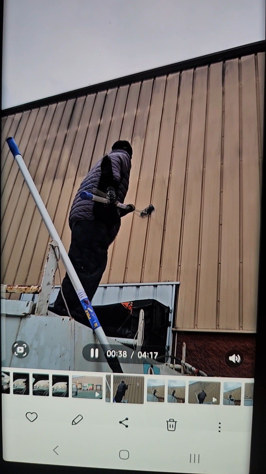A person on a ladder uses a long-handled paint roller to apply paint to a tall, vertical metal-sided building.