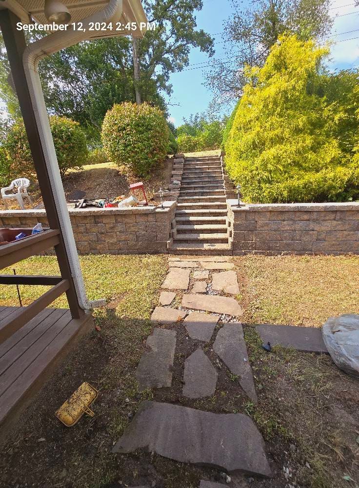 A stone pathway leads toward a set of stone steps and a short retaining wall in a sunlit backyard with green landscaping.