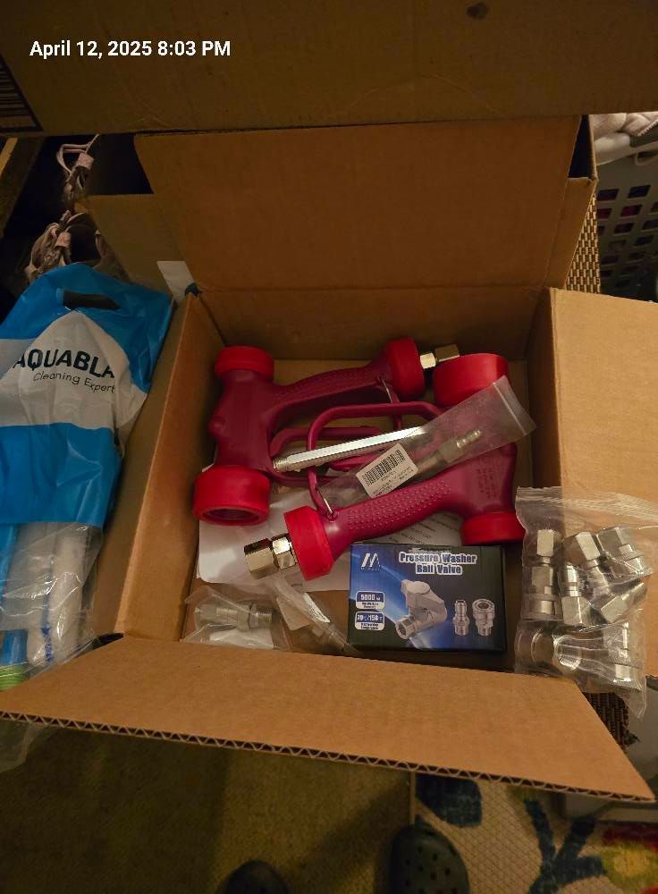 An open cardboard box containing two red high-pressure spray guns, various metal fittings, and a bagged accessory kit.