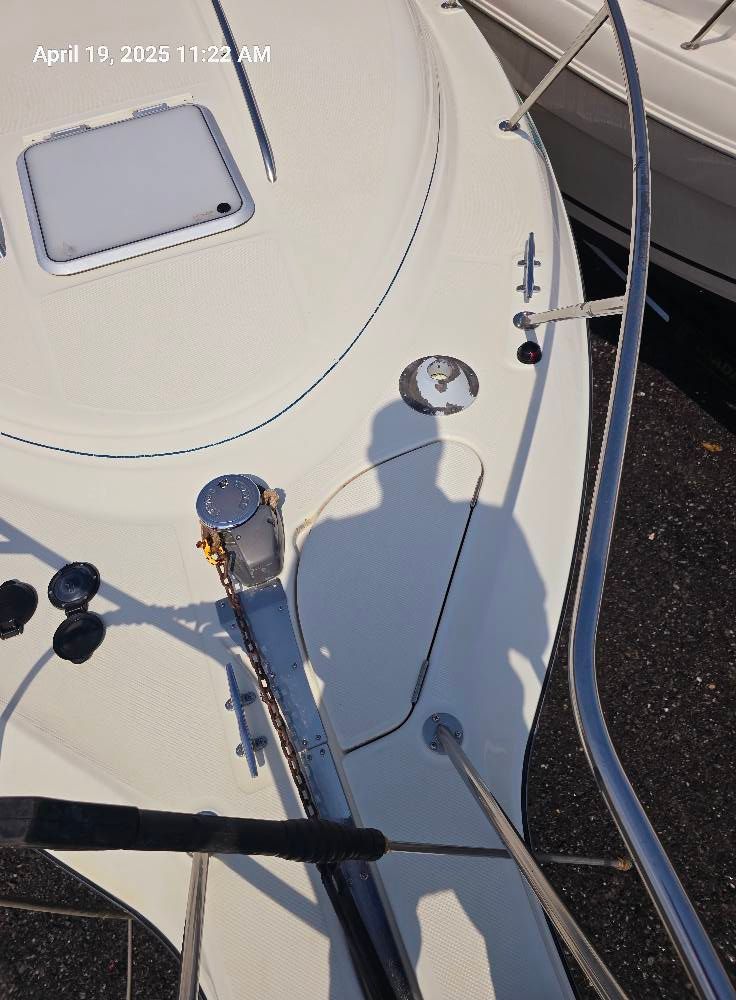 A high-angle view of a boat bow, featuring an anchor windlass, a deck hatch, and a person’s shadow cast onto the deck.