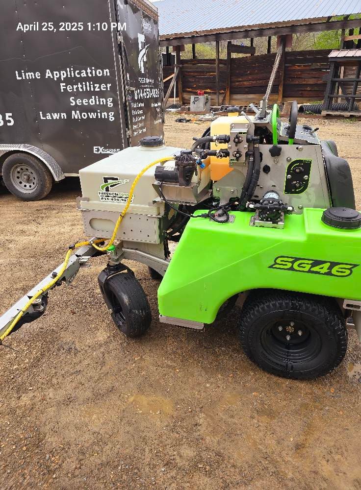A lime-green Turfco SG46 spreader-sprayer machine parked next to a utility trailer with lawn care service information.