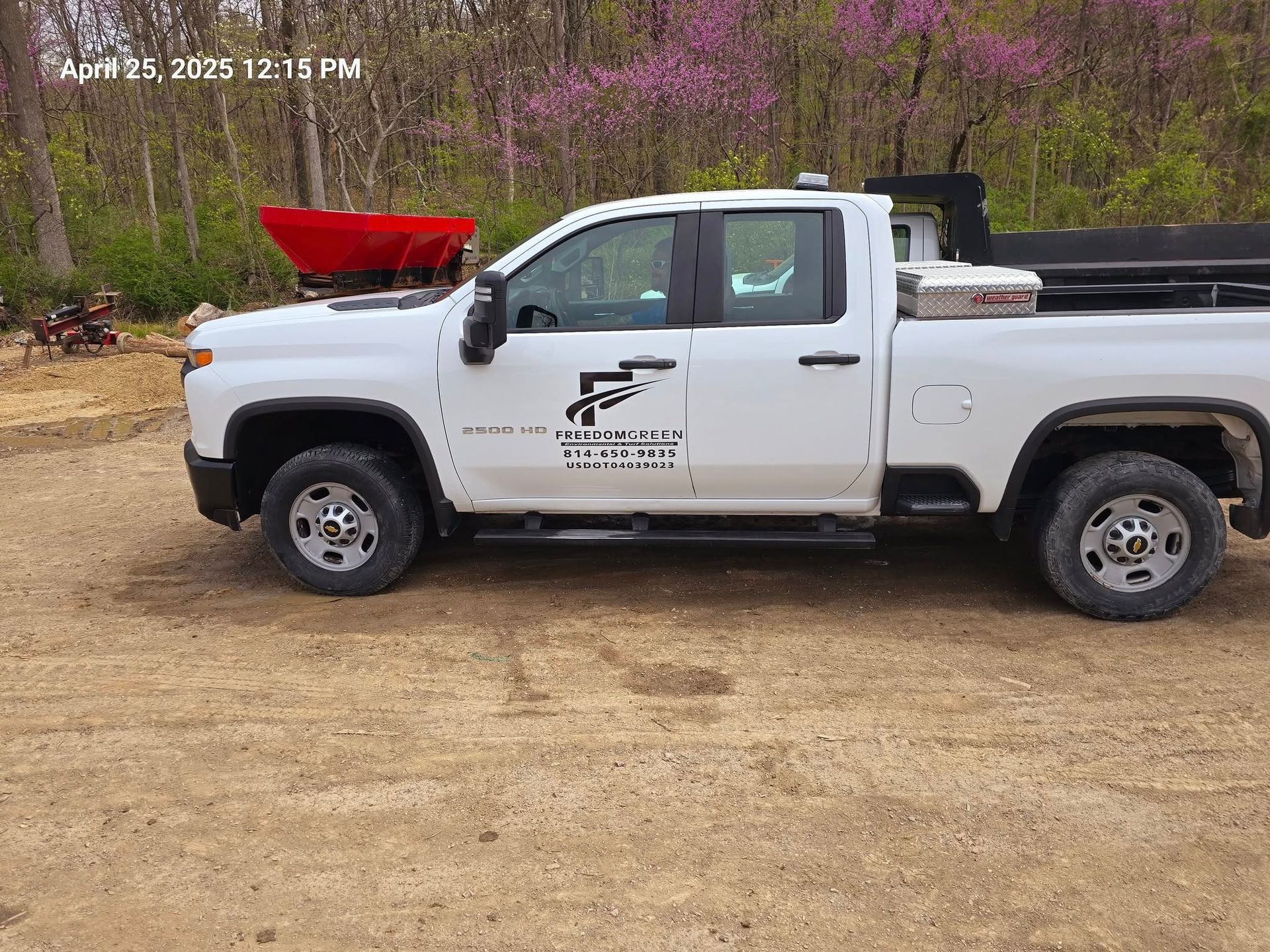 A white Chevrolet work truck parked on a dirt lot, featuring a company logo on the door and a red spreader in the background.
