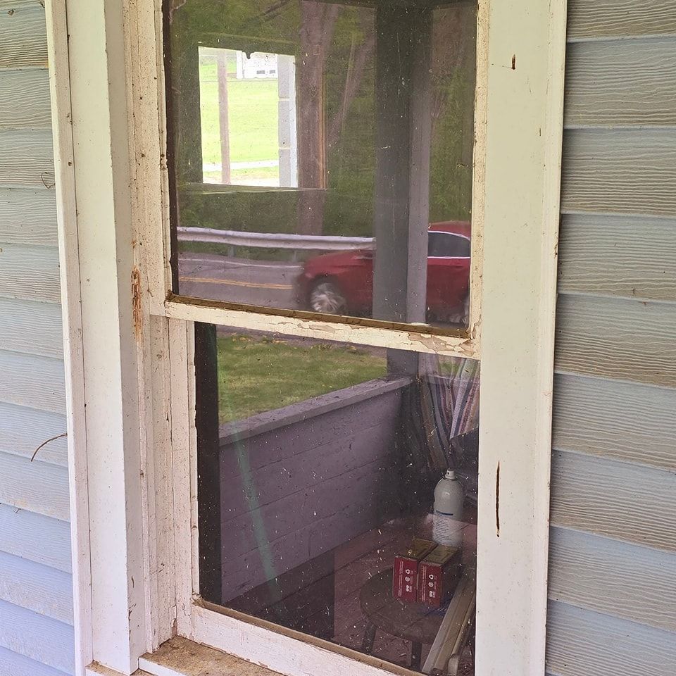 A weathered white window frame set in light blue siding, reflecting a red car and a porch in the glass.