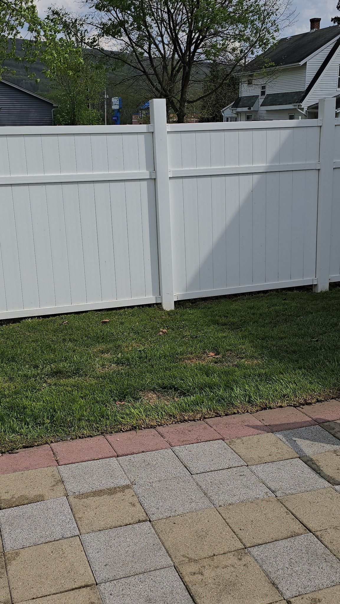 A white vinyl privacy fence separates a grassy backyard from a patio made of light and dark square stone pavers.