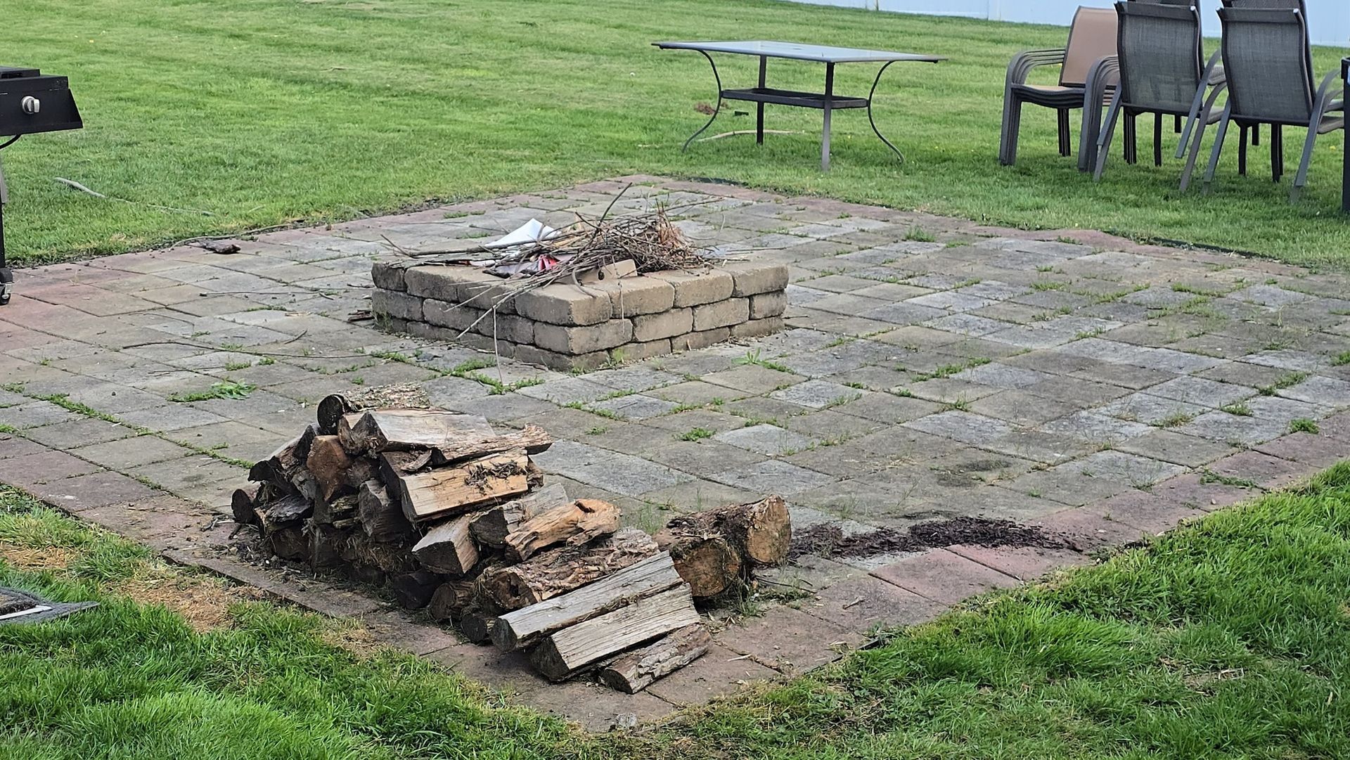 A brick patio features a central stone fire pit, a stack of firewood in the foreground, and outdoor furniture nearby.