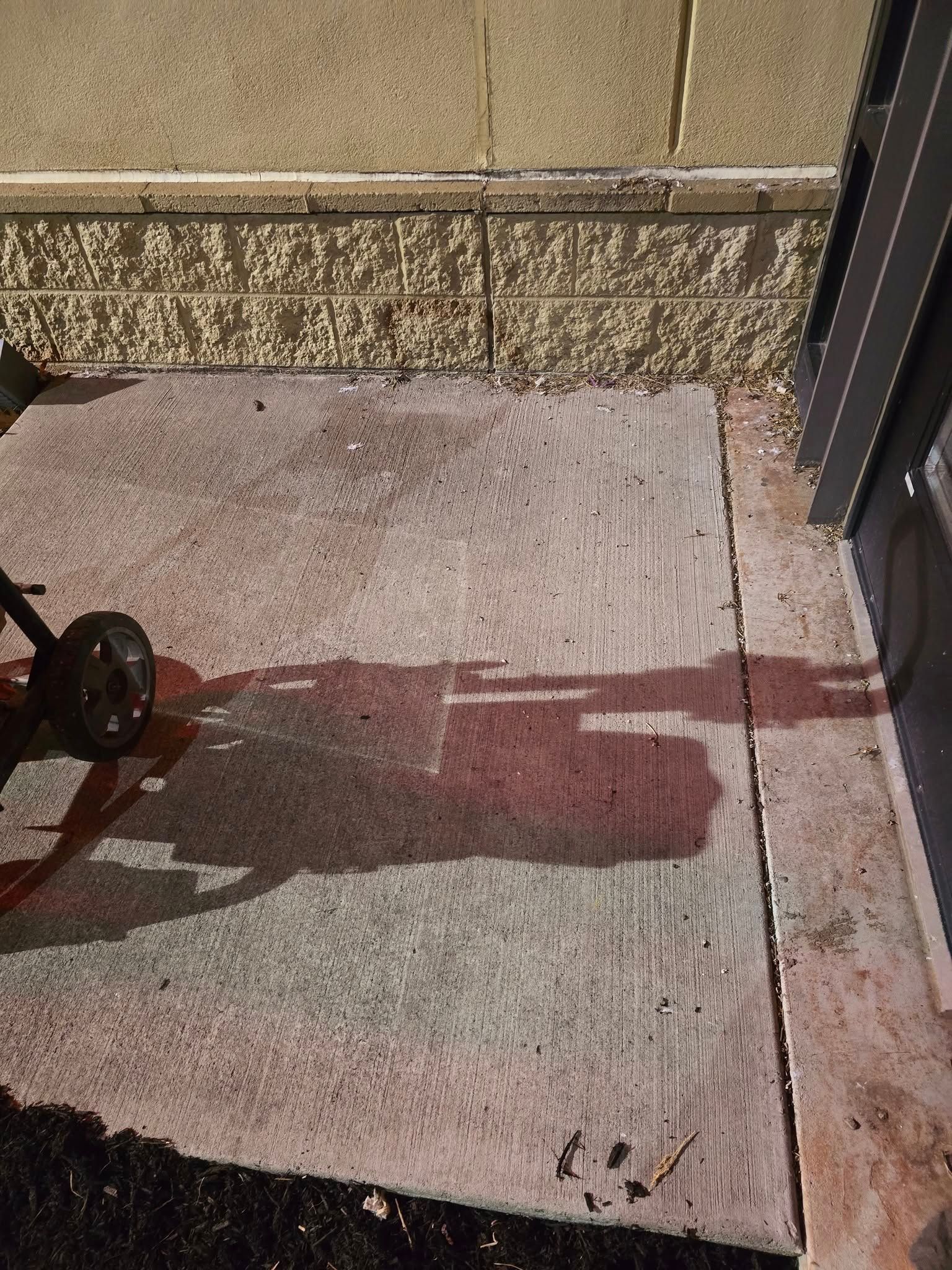 A lawn mower's shadow cast on a concrete patio next to a beige building wall.