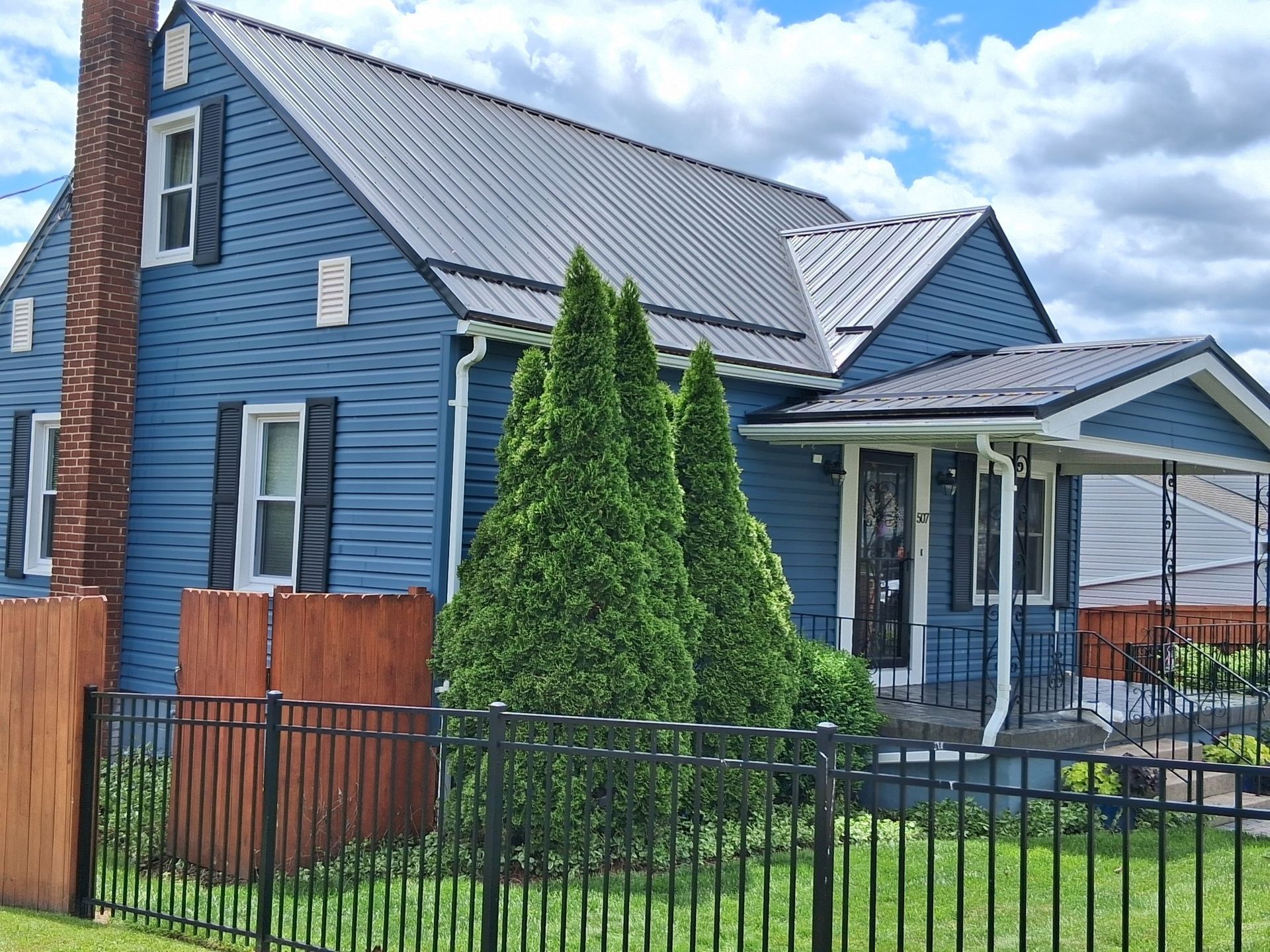 A blue house with a gray metal roof, a brick chimney, and black shutters, behind a black metal fence and green trees.
