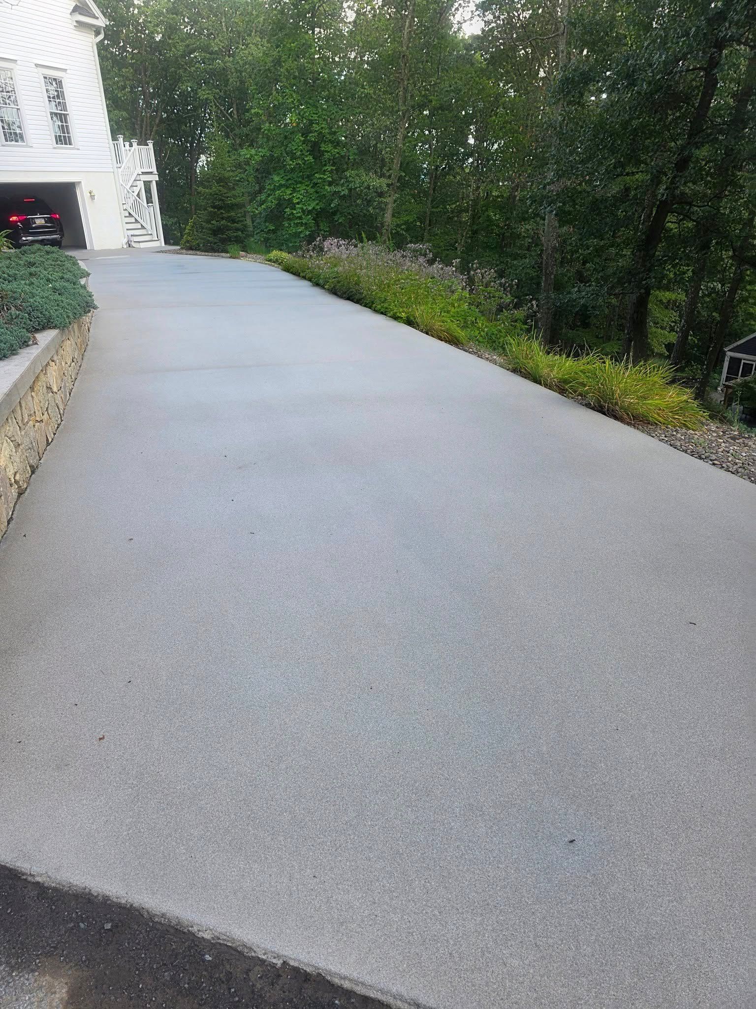 A smooth concrete driveway leads toward a white house surrounded by green trees and a low stone retaining wall.