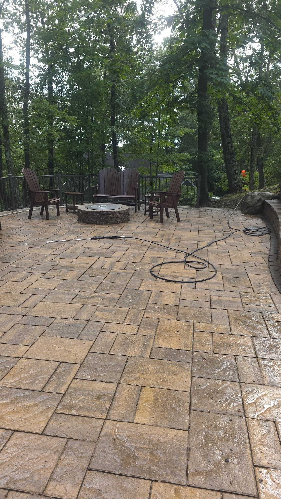 A stone patio with a fire pit, three chairs, and a coiled garden hose, situated in a backyard surrounded by forest trees.