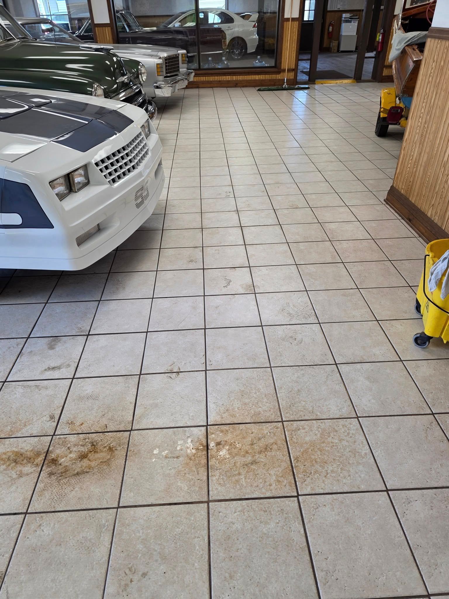 A white car and a green car parked inside a garage with light-colored tile flooring and some dirt patches.