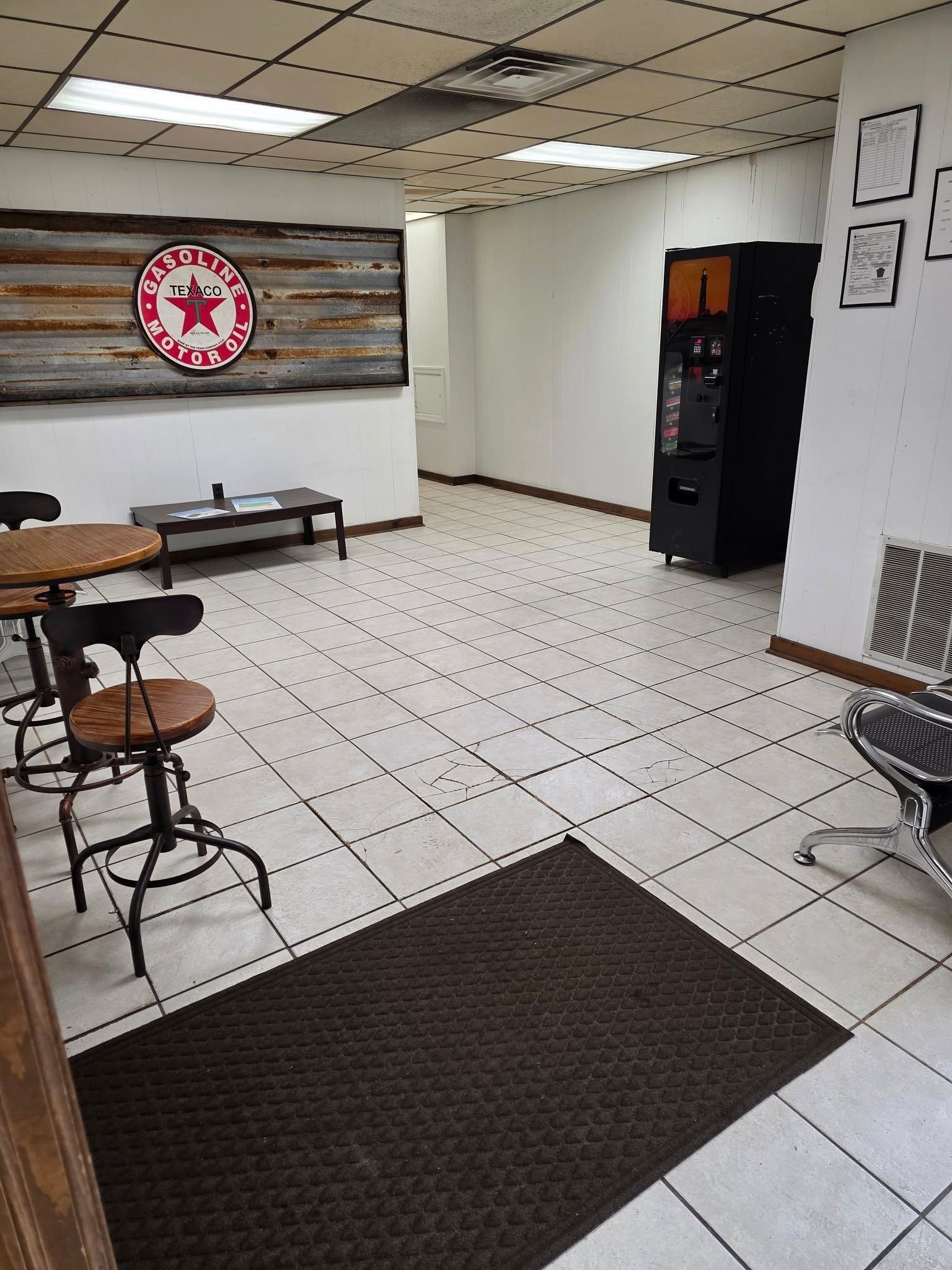 A waiting area featuring a tile floor, a rustic wall sign, a small table, bar-style chairs, and a black vending machine.
