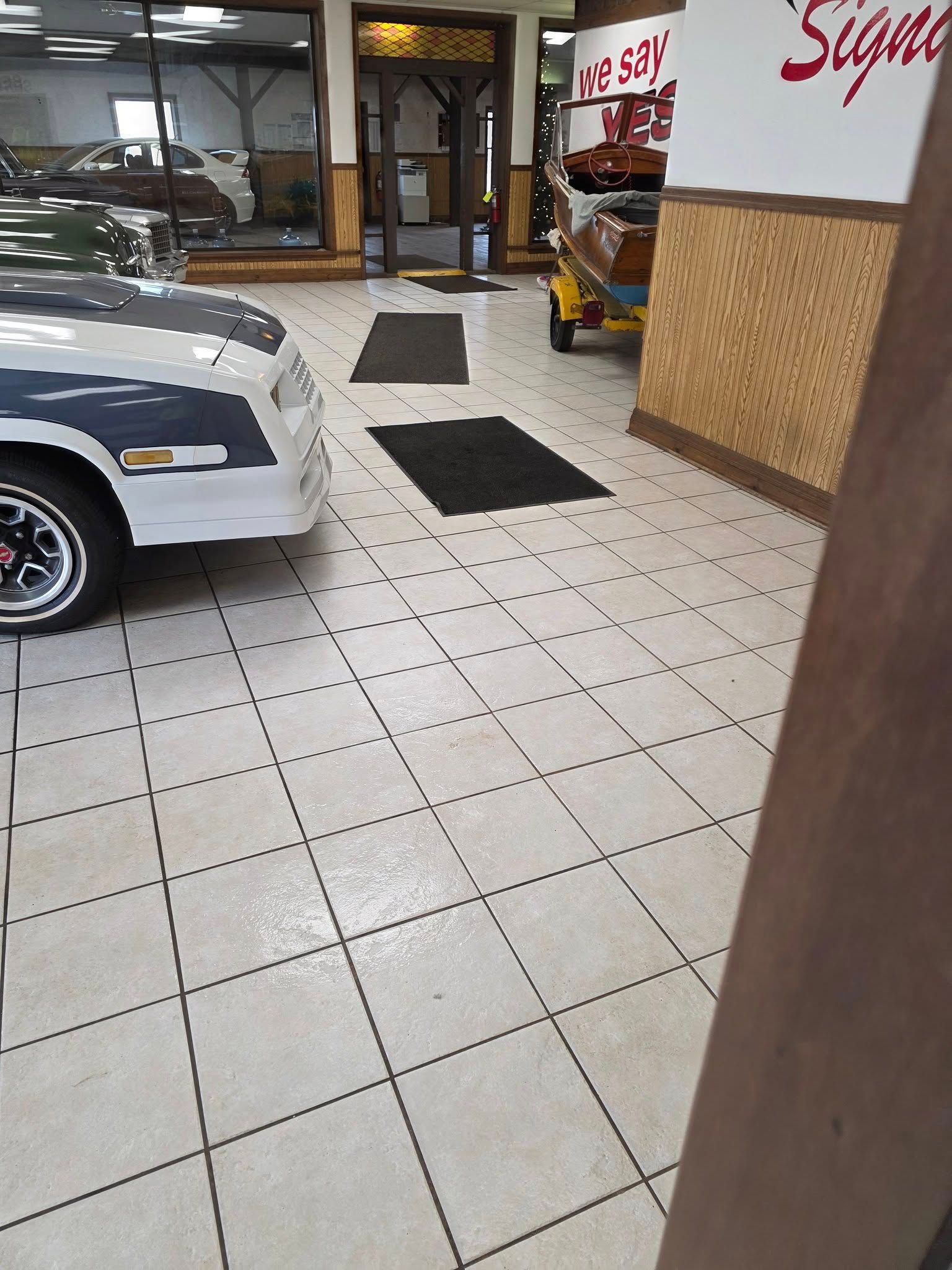 A showroom with a tiled floor, two black floor mats, a parked classic white car, and a boat on a trailer in the background.