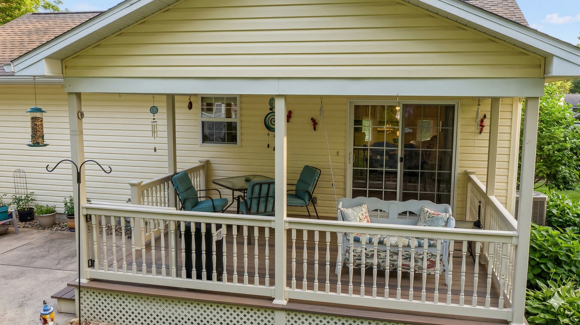 A yellow-sided house featuring a spacious covered wooden deck with outdoor seating and a railing, overlooking a patio.