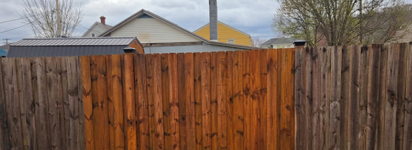 A section of a wooden fence shows a stark contrast between weathered grey boards and freshly stained, warm-toned wood.