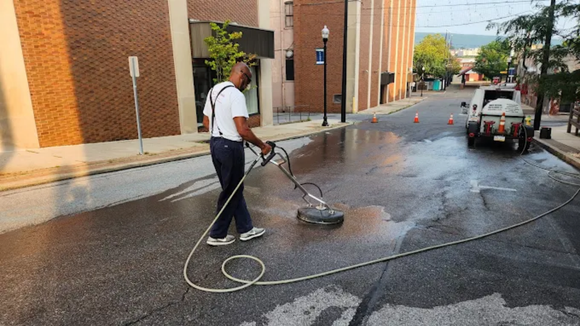A worker uses a surface cleaner to power wash a paved street in an urban area.