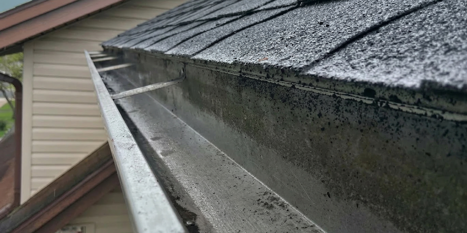 A view of a metal rain gutter mounted on the edge of a roof with asphalt shingles, showing metal support straps.