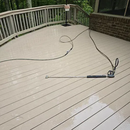 A pressure washer wand rests on a clean, light-colored wooden deck surrounded by railings and a brick wall.