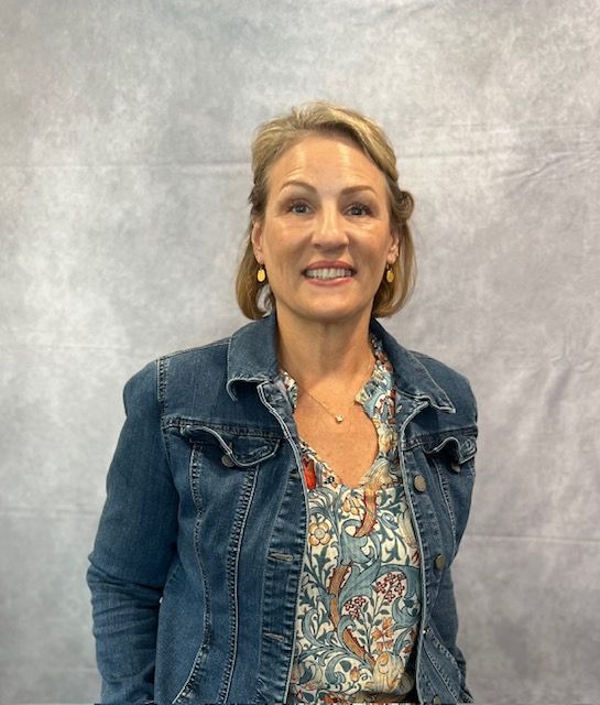 A woman wearing a denim jacket and a floral shirt is smiling for the camera.